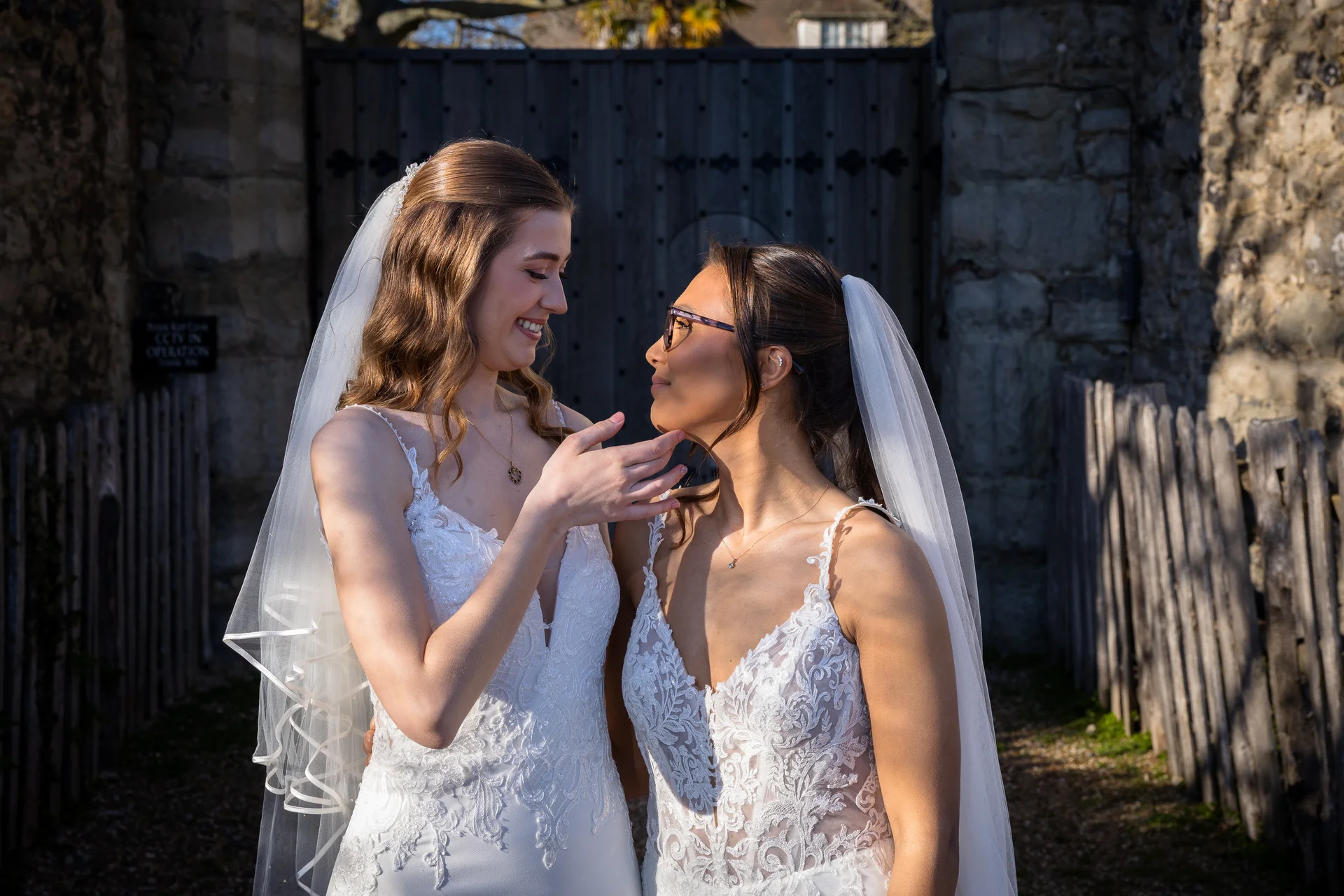 Two smiling brides in matching lace wedding dresses and veils stand outside a stone courtyard, sharing a tender moment in the golden sunlight.
