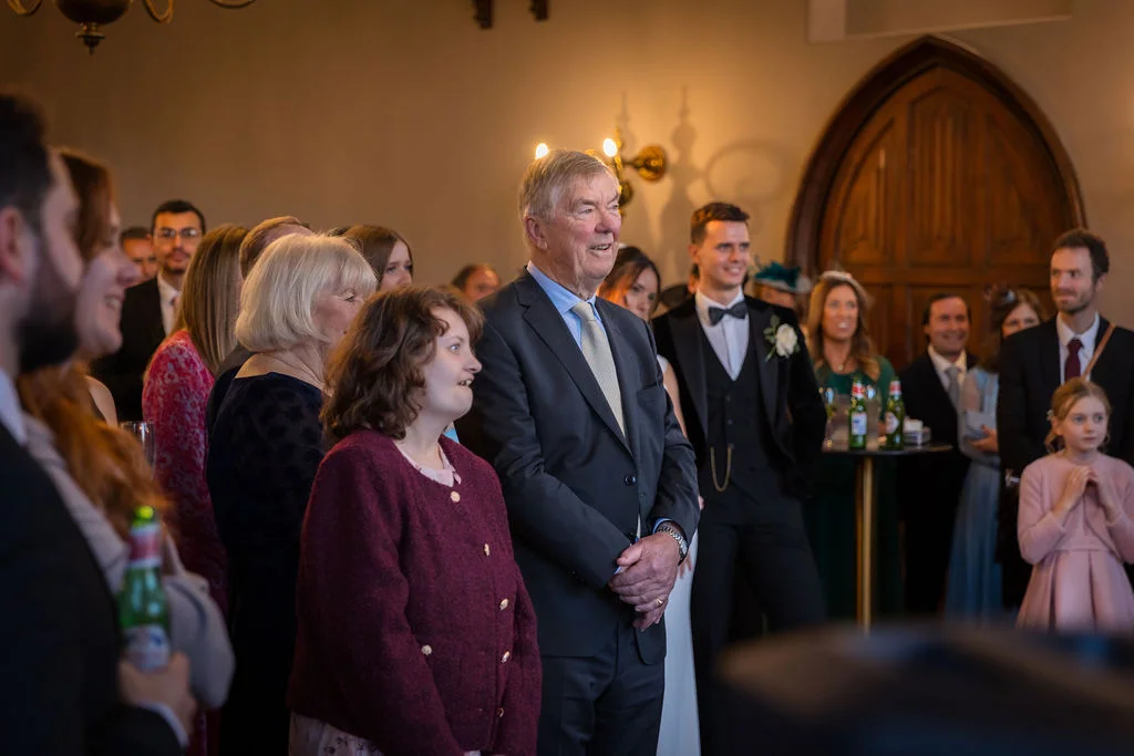 Wedding guests gathered indoors, smiling and laughing together during speeches in a venue with a stone archway.
