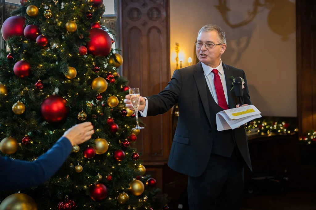 A smiling guest delivers a speech while holding a glass of champagne next to a large, festively decorated Christmas tree during a winter wedding reception.