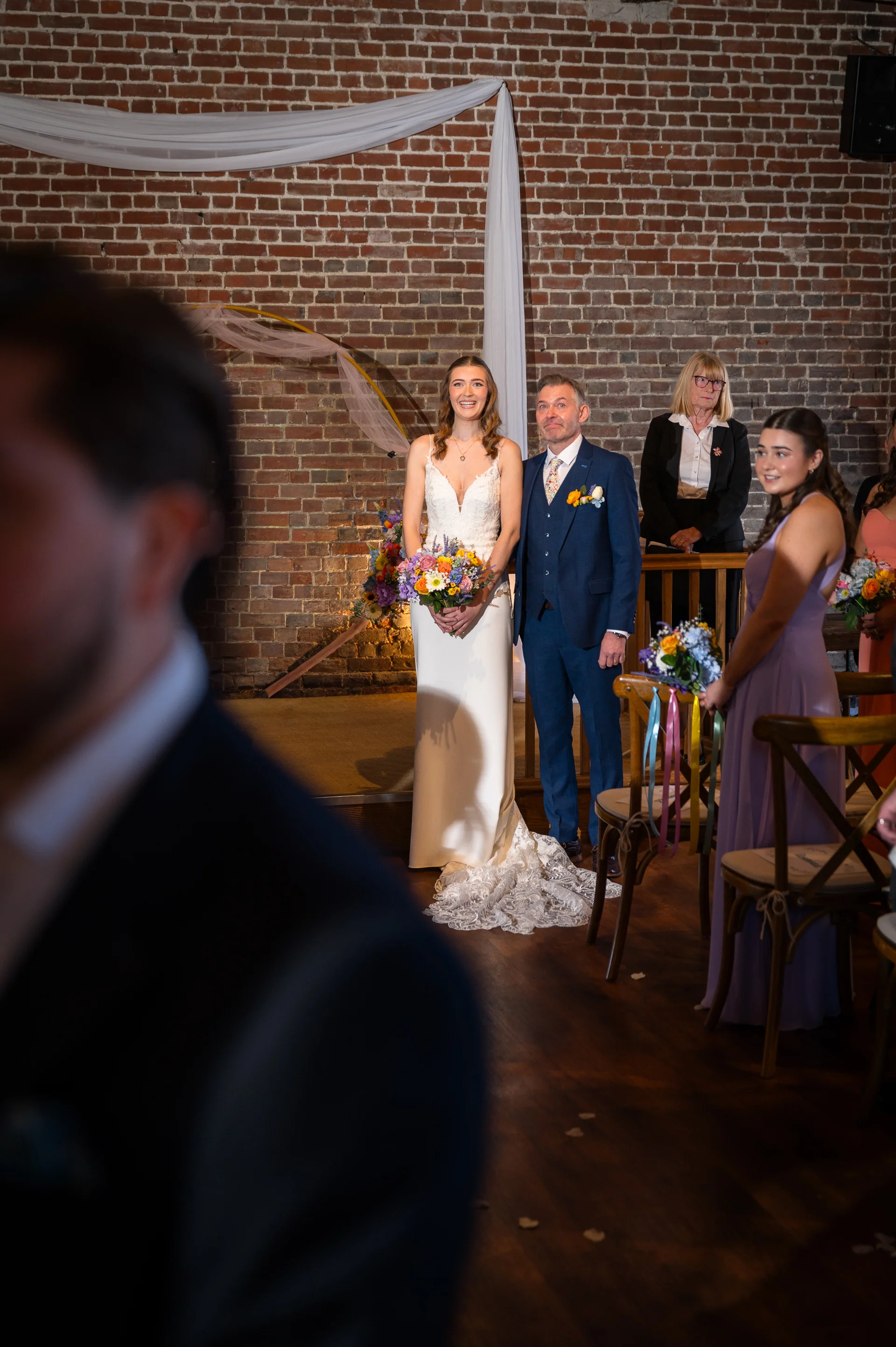A bride in a fitted V-neck gown and groom in a blue suit stand together at the altar inside a rustic stone barn venue during their wedding ceremony.