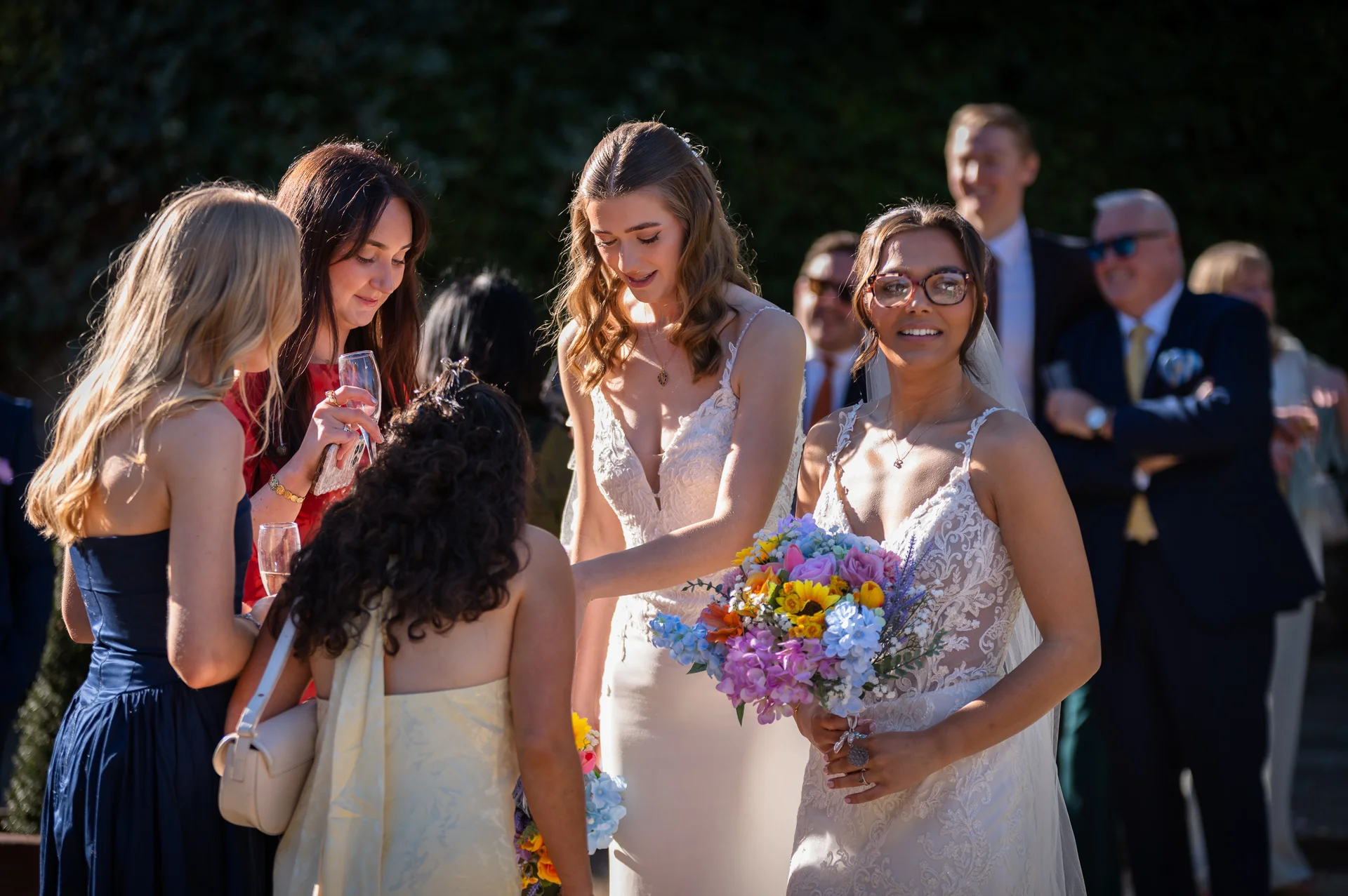 Two smiling brides stand outdoors surrounded by wedding guests during a sunlit drinks reception, one holding a colourful bouquet of flowers.