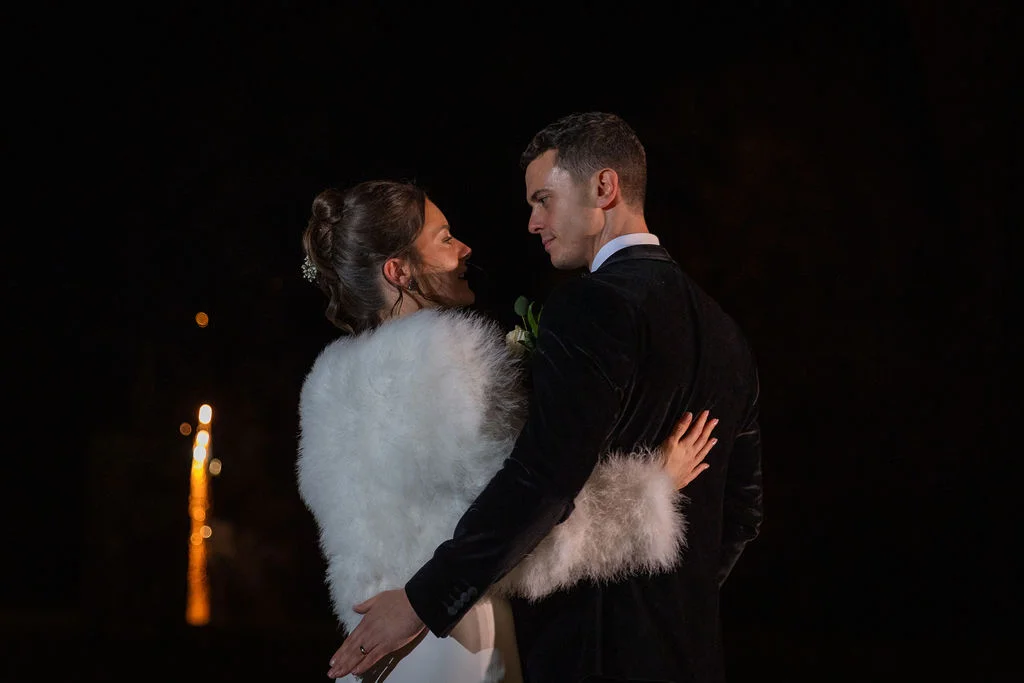 A newlywed couple embraces outdoors at night during winter, with the bride wearing a white faux fur stole and the groom in a dark velvet tuxedo.