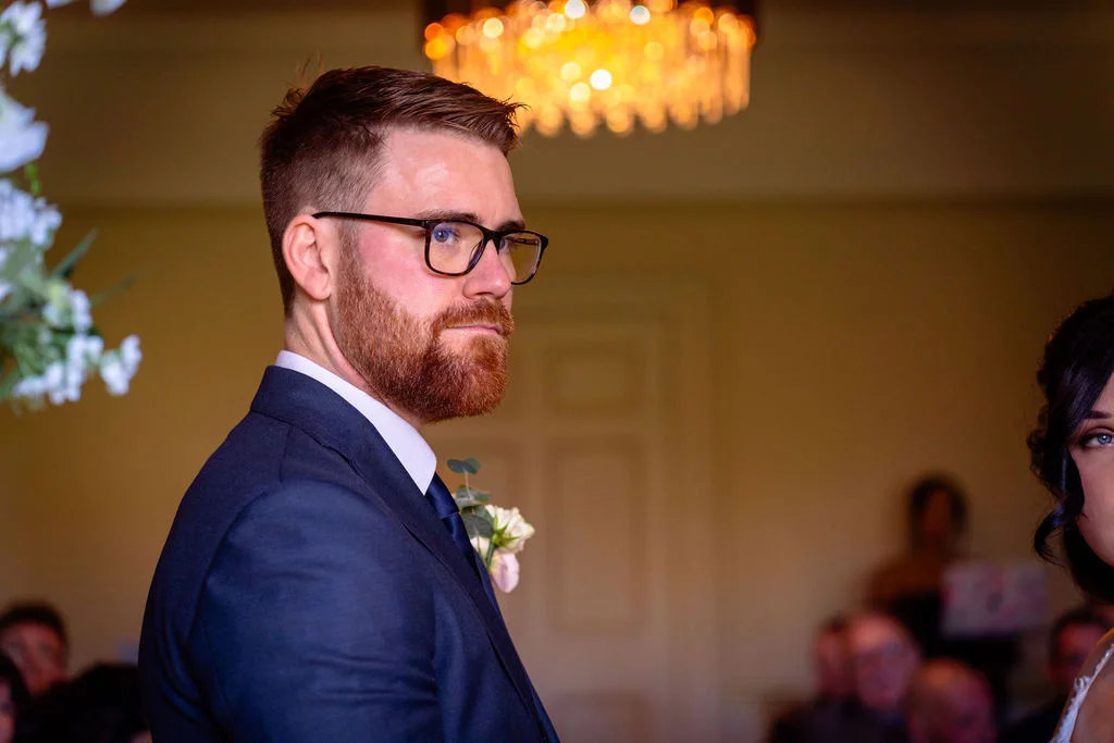 A groom in a navy suit with a boutonniere stands waiting at the altar during a wedding ceremony, illuminated by soft side light.