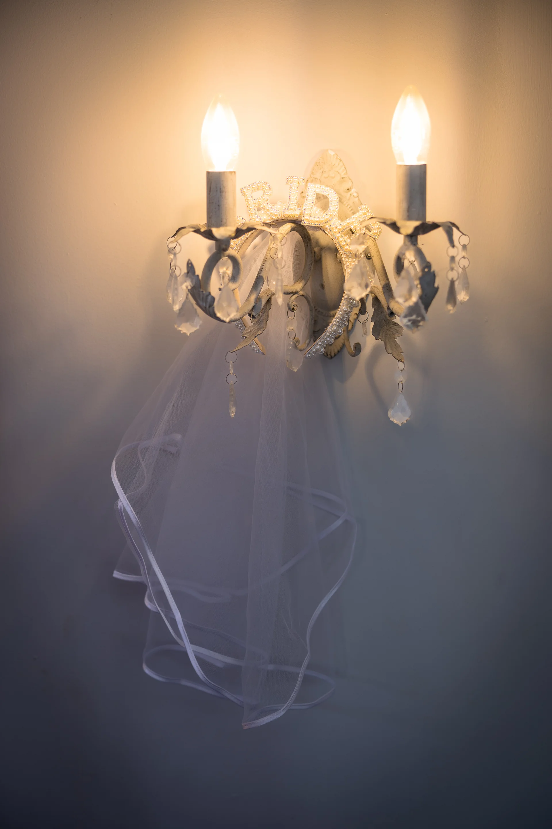 A delicate white bridal veil hangs gracefully over an ornate wall sconce featuring lit candles and crystal droplets.
