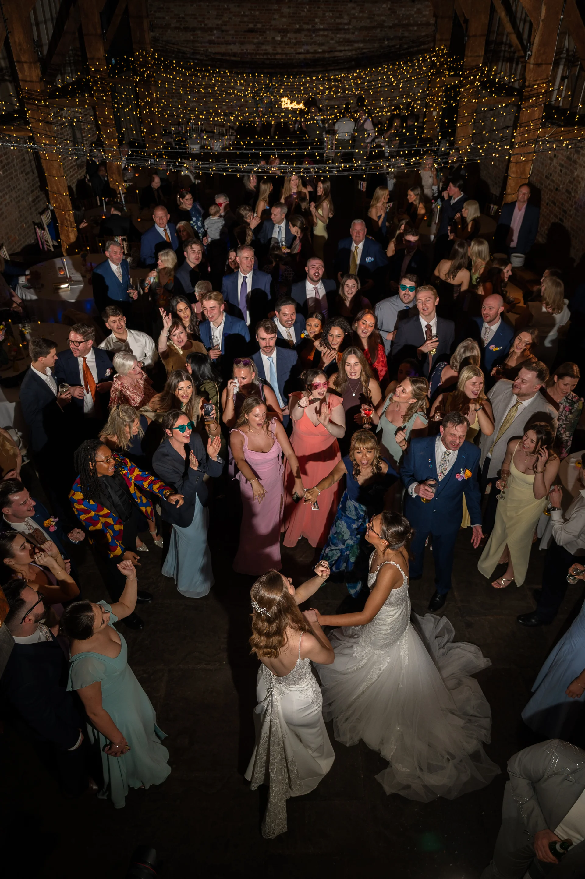 An elevated view captures a joyful same sex couple dancing together surrounded by a large crowd of cheering wedding guests inside a rustic barn illuminated by a canopy of fairy lights.