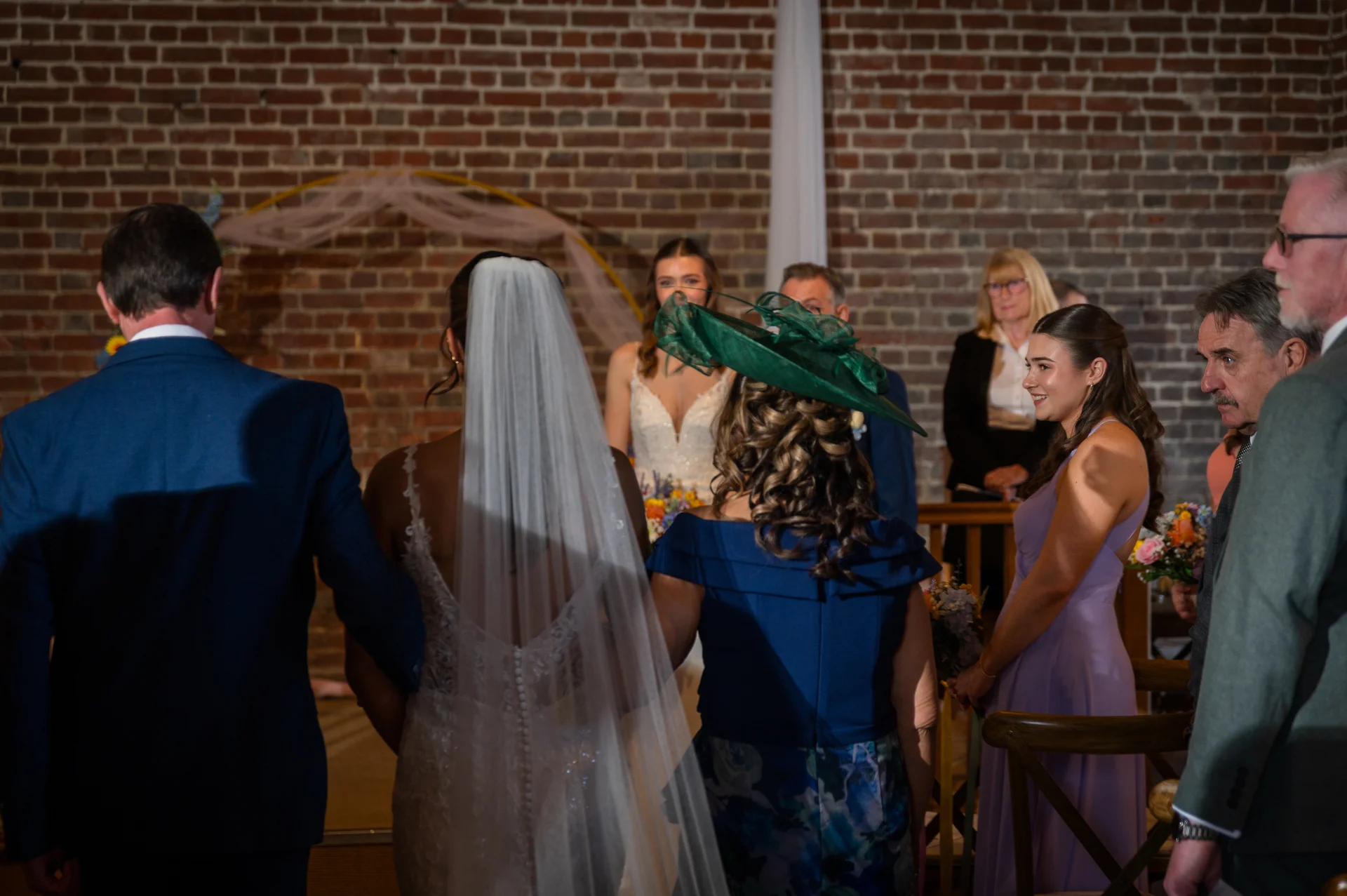The bride and groom walk down the aisle together during their ceremony, framed by guests and a rustic exposed brick wall.