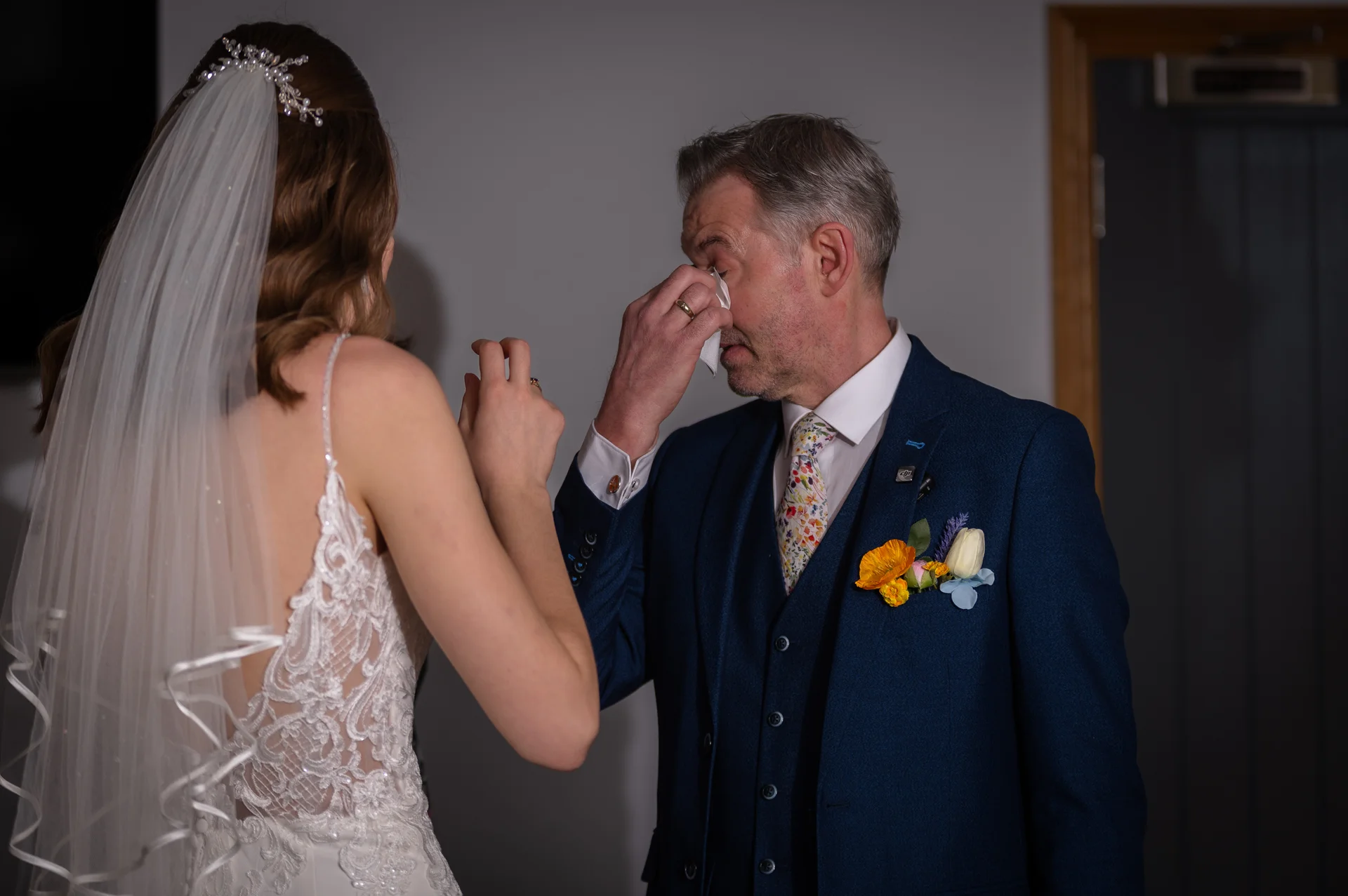 An emotional father of the bride wipes away tears with a tissue while wearing a blue three-piece suit during a tender moment with his daughter.