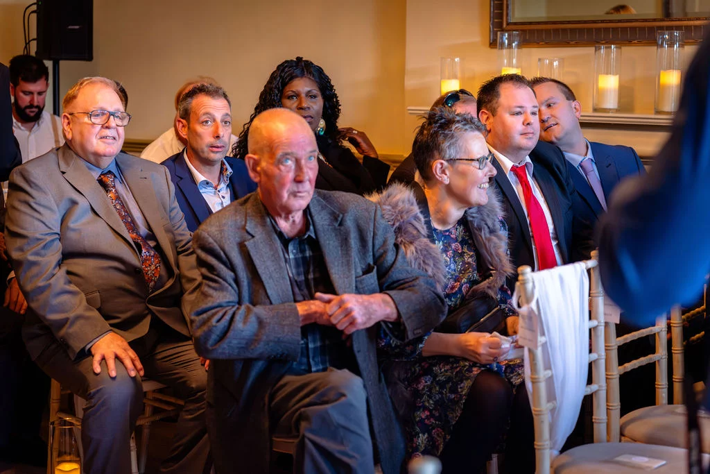 A candid group of wedding guests seated and laughing together during a speech inside a venue.