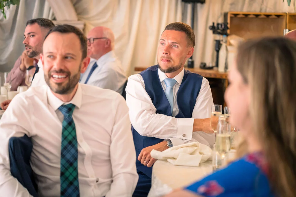 Groomsmen and guests sitting together at a wedding reception listening attentively to speeches and sharing a joyful moment.