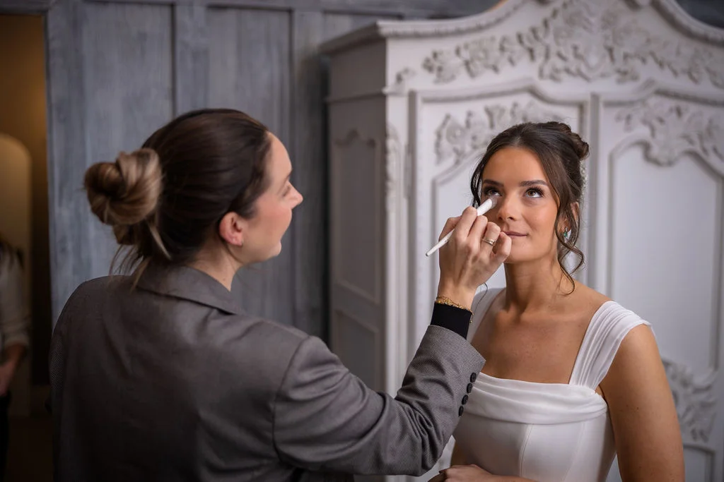 A bride receiving delicate eye makeup application from a stylist in a grey blazer during morning bridal preparations.
