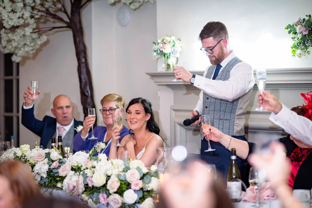 A groom delivers a speech holding a microphone and raising a champagne flute while seated guests at the top table smile and toast.