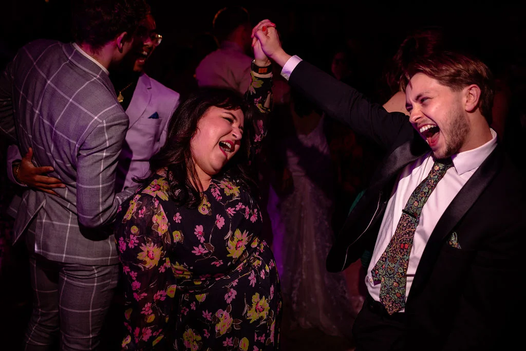 Wedding guests laugh and dance enthusiastically together on a dimly lit evening dance floor with energetic movement.