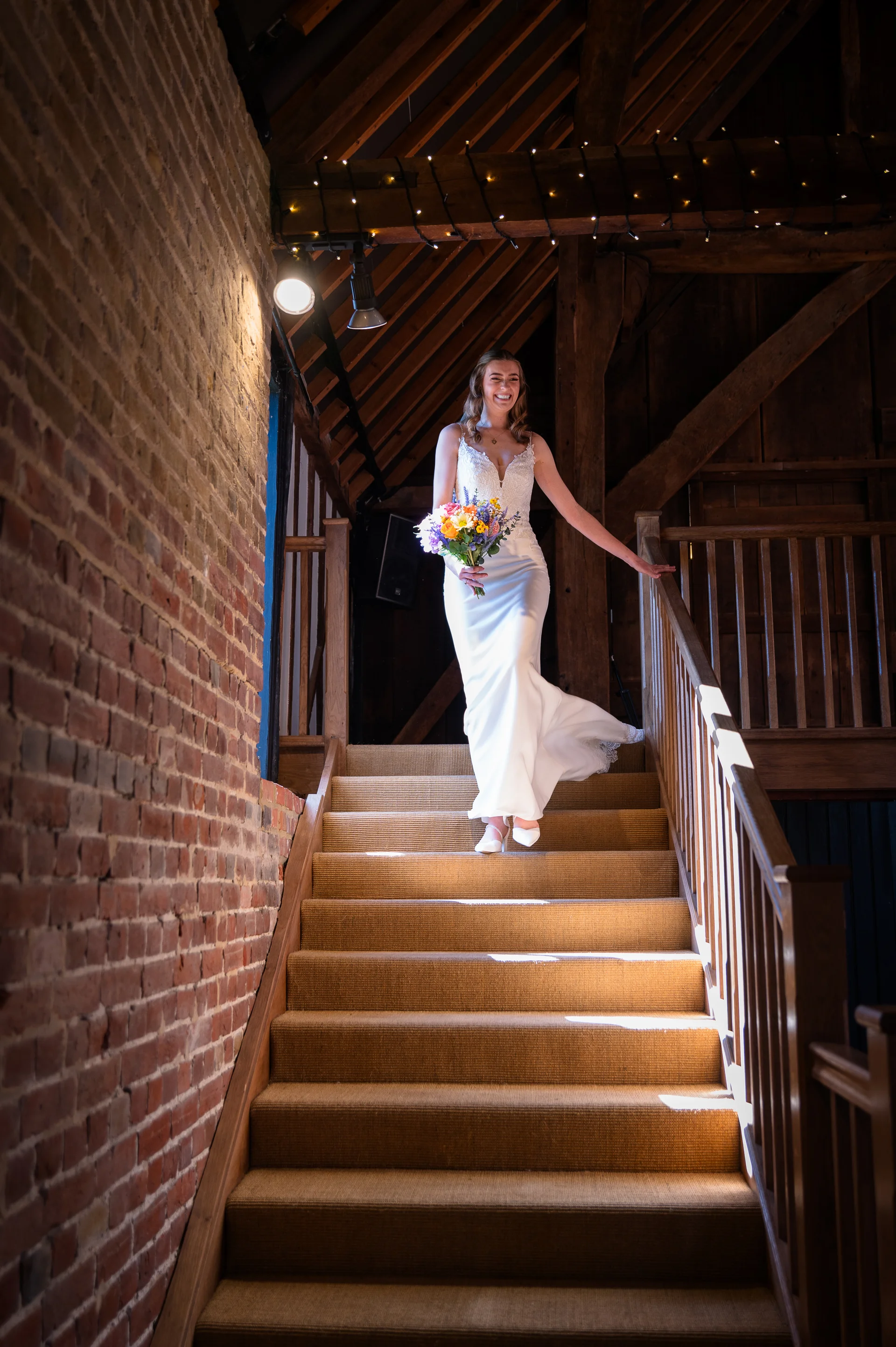 A joyful bride descends a wooden staircase in a rustic barn venue, holding a colourful hand tied bouquet and wearing a sleek crepe gown with a v neckline.