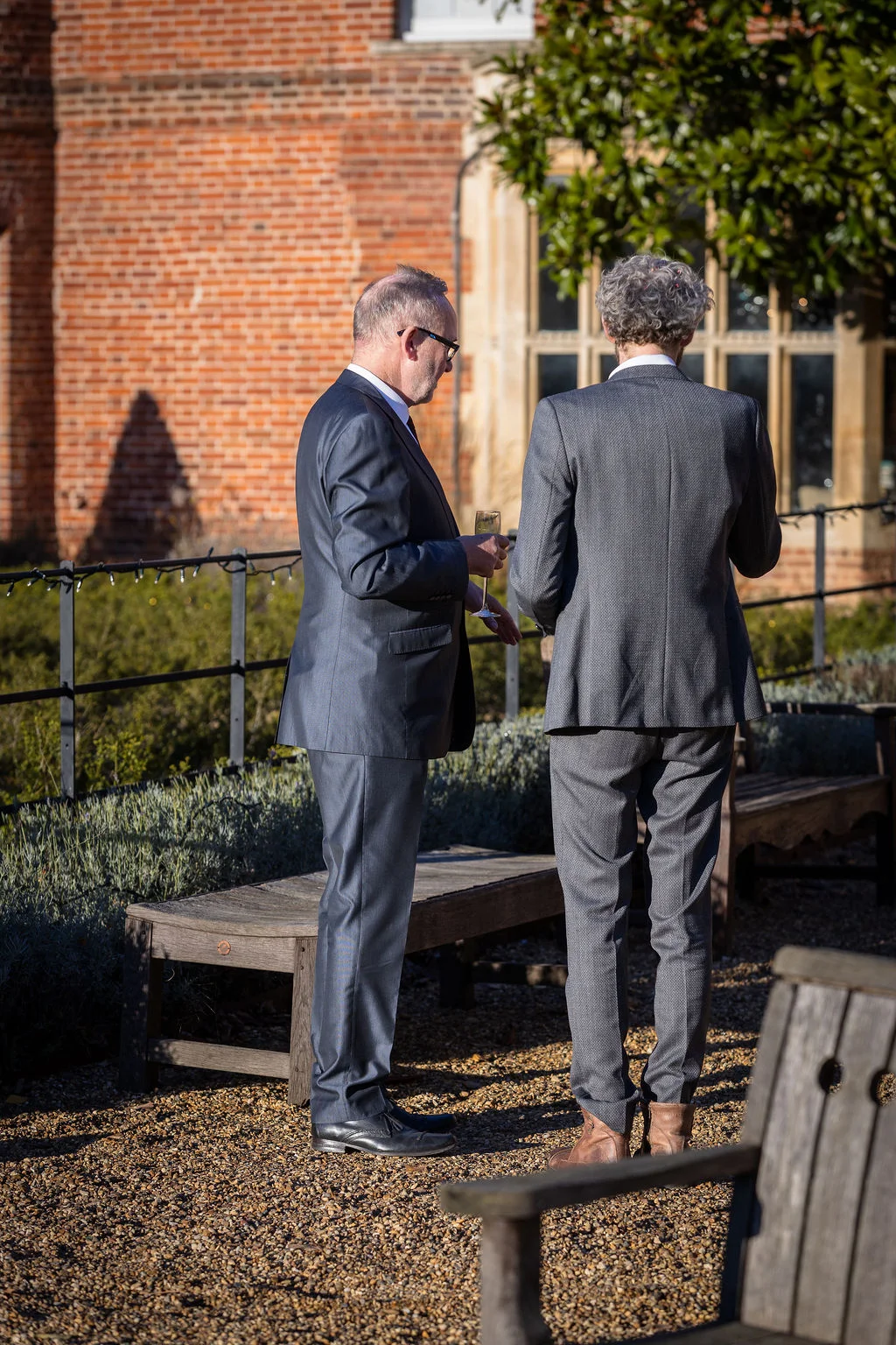 Two male guests in smart suits chat outside in a gravel courtyard during a wedding drinks reception.
