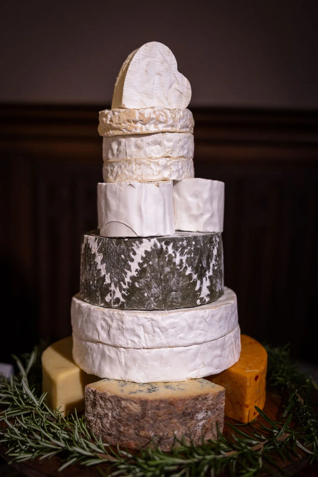 A magnificent tiered wedding cheese tower featuring a variety of artisanal rounds displayed with fresh rosemary sprigs at the base.