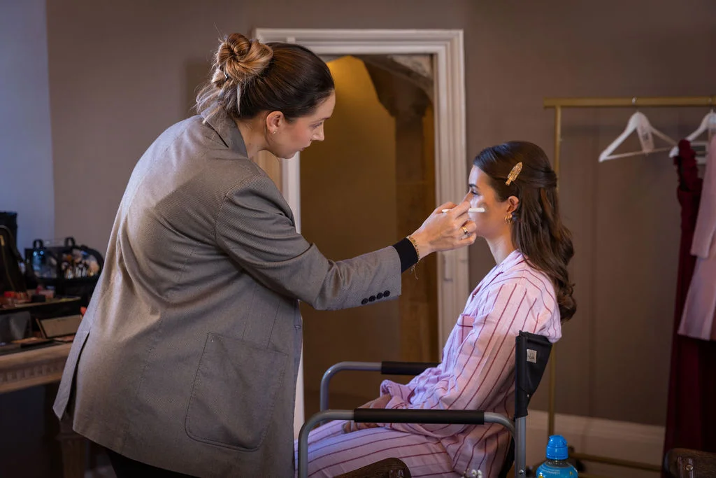 A makeup artist applies finishing touches to a bride sitting in a director's chair while wearing striped pink pyjamas during morning bridal prep.