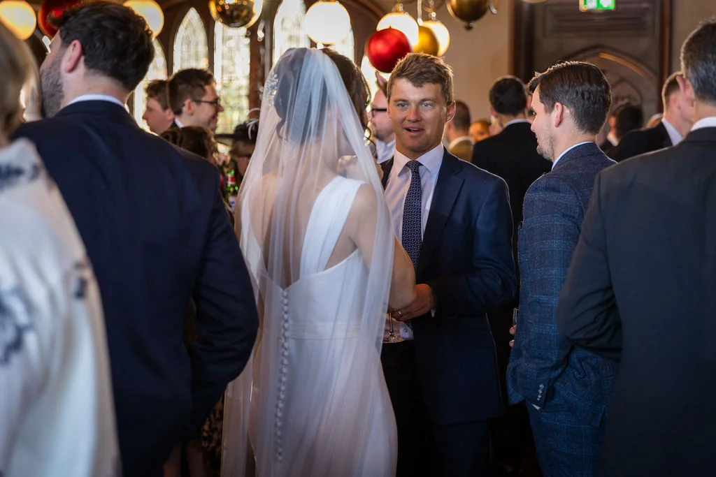 A bride in a button-back gown with a chapel veil and a groom in a navy suit mingle with guests during a wedding reception.