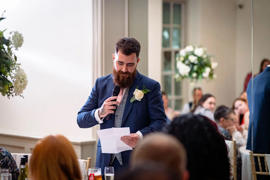 A bearded groom in a navy suit and tweed waistcoat delivers a heartfelt speech holding a microphone and notes during the wedding breakfast.