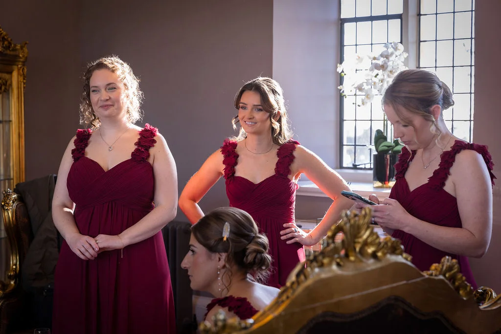 Three bridesmaids in matching burgundy chiffon dresses with ruched bodices stand near a window while a fourth bridesmaid is seated in the foreground.