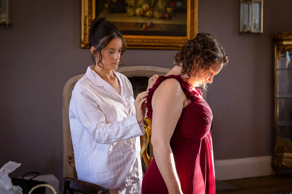 A bridesmaid wearing a white pyjama shirt helps another bridesmaid into her pleated burgundy dress inside a classically styled country house room illuminated by soft window light.