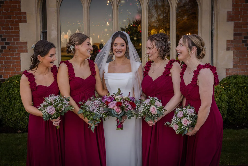 A smiling bride holding a mixed floral bouquet stands outdoors in front of a stone archway with four bridesmaids wearing matching burgundy dresses.
