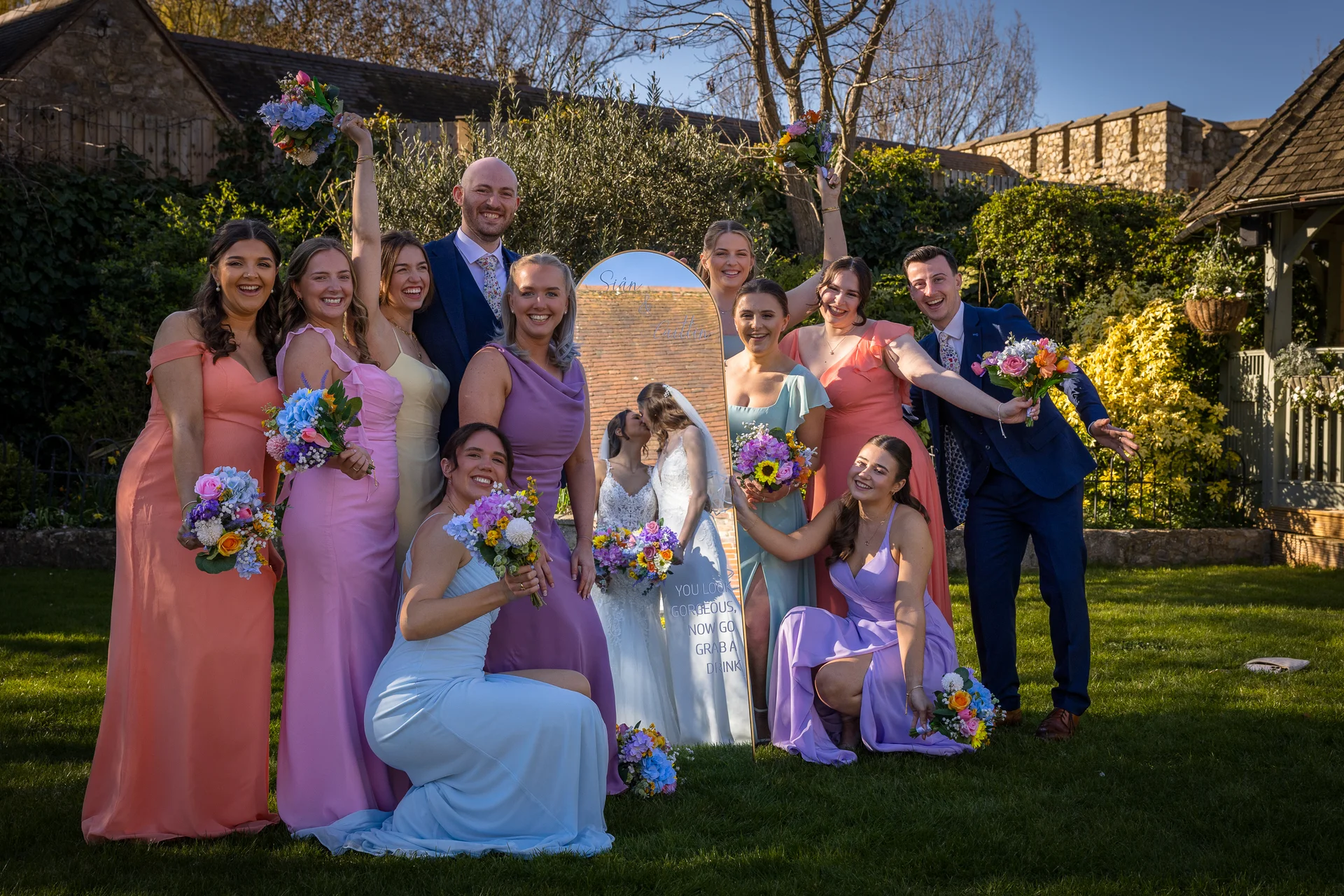 A joyful wedding party in pastel dresses and blue suits poses outdoors on a lawn, reflected in a decorative mirror sign.