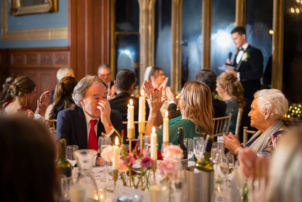 Wedding guests seated at banquet tables during the speeches, with a man wiping a tear and others laughing and applauding in a stately venue.