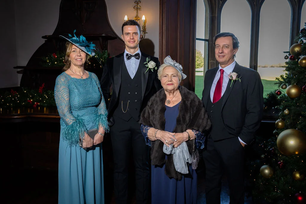 A formal family group portrait featuring a groom in a black tuxedo alongside older family members, including a woman in a teal dress and fascinator and guests next to a decorated Christmas tree inside a venue.