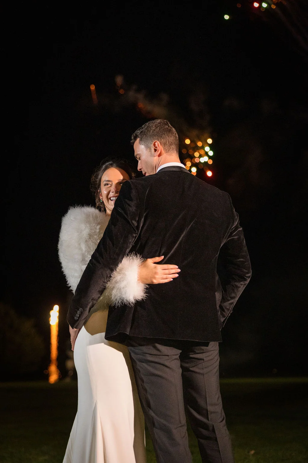 A newlywed couple embraces outdoors at night under a display of fireworks, with the bride wearing a sleek white dress featuring feathery cuffs and the groom in a black velvet tuxedo.