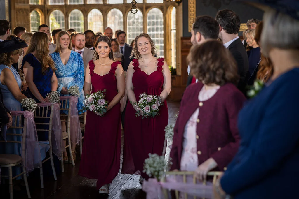 Two smiling bridesmaids in matching burgundy gowns walk down the aisle holding floral bouquets during a wedding ceremony inside a historic country house.