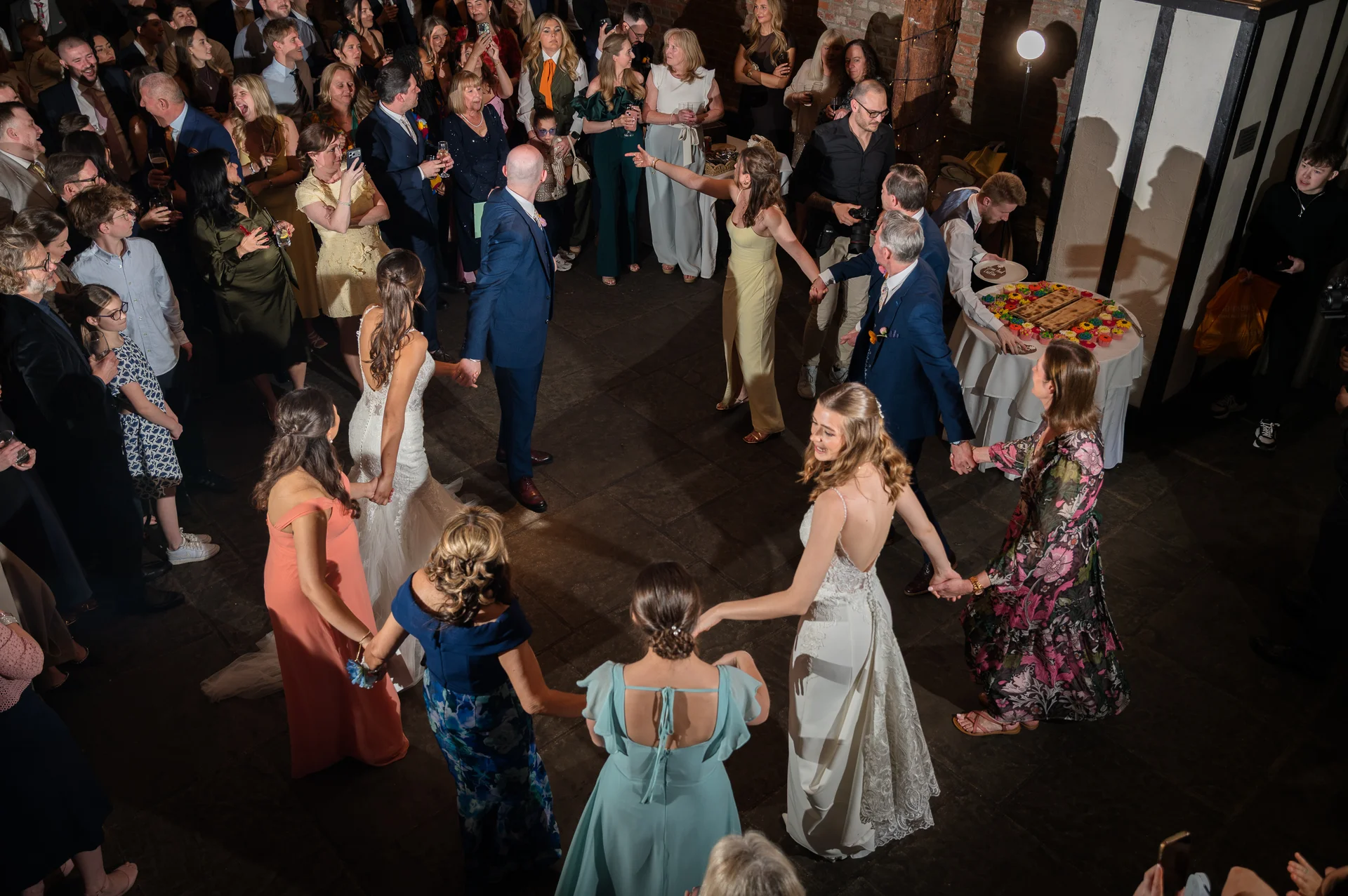 An overhead view captures wedding guests and the newly married couple holding hands to form a large circle dancing on a stone floor during the evening reception.