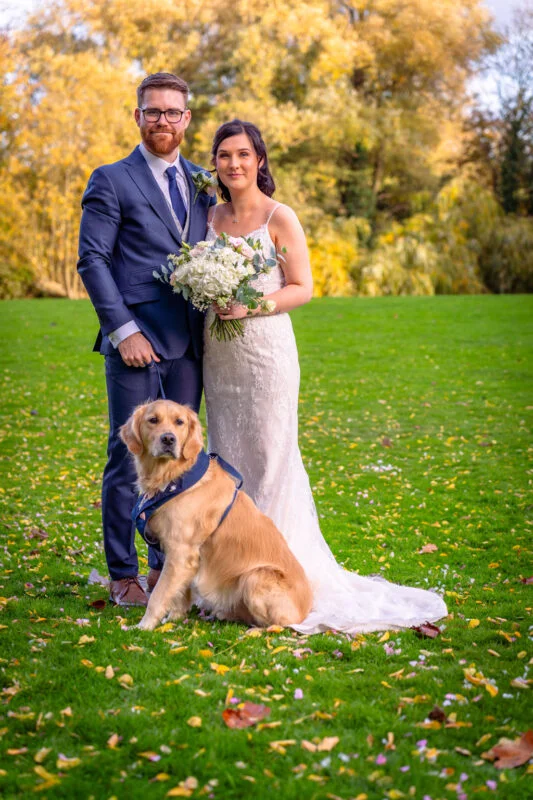A bride and groom pose on a green lawn with their golden retriever in an autumn garden setting.