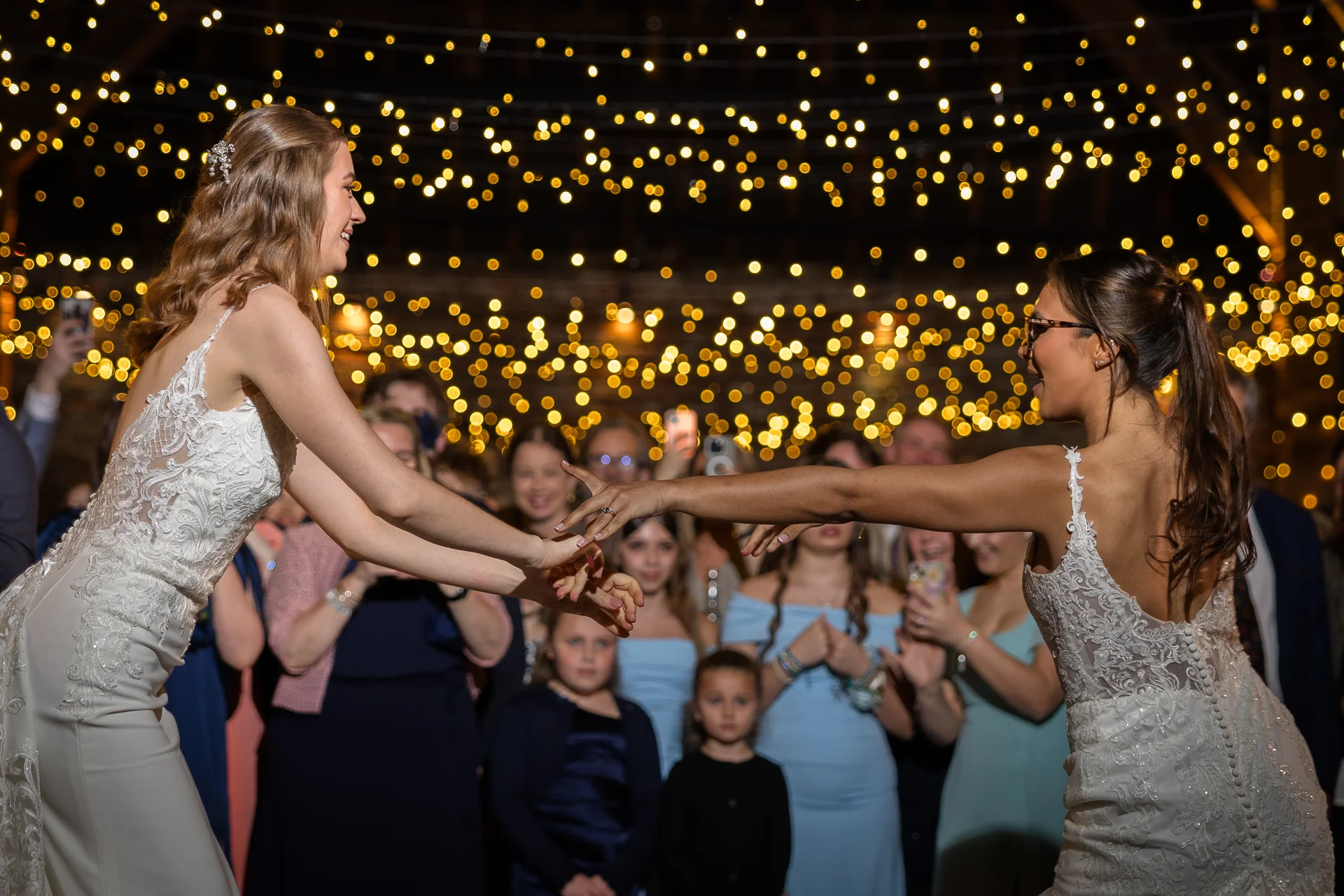 Two smiling brides in lace wedding dresses hold hands and dance together during their evening reception beneath a warm canopy of fairy lights.
