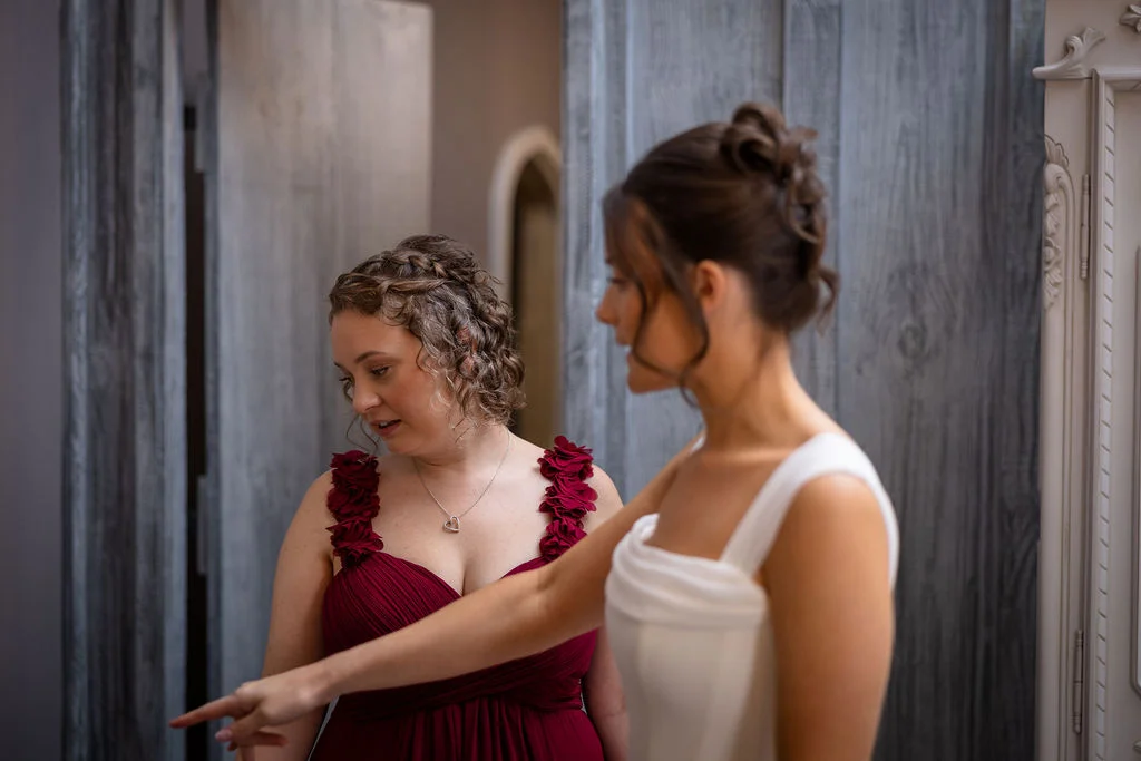 A bride in a modern white gown points out a detail to her bridesmaid, who wears a burgundy dress with floral strap details during morning preparations.
