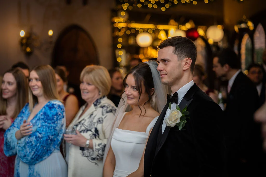 Newlywed couple smiling proudly alongside wedding guests in an indoor venue illuminated by warm fairy lights.