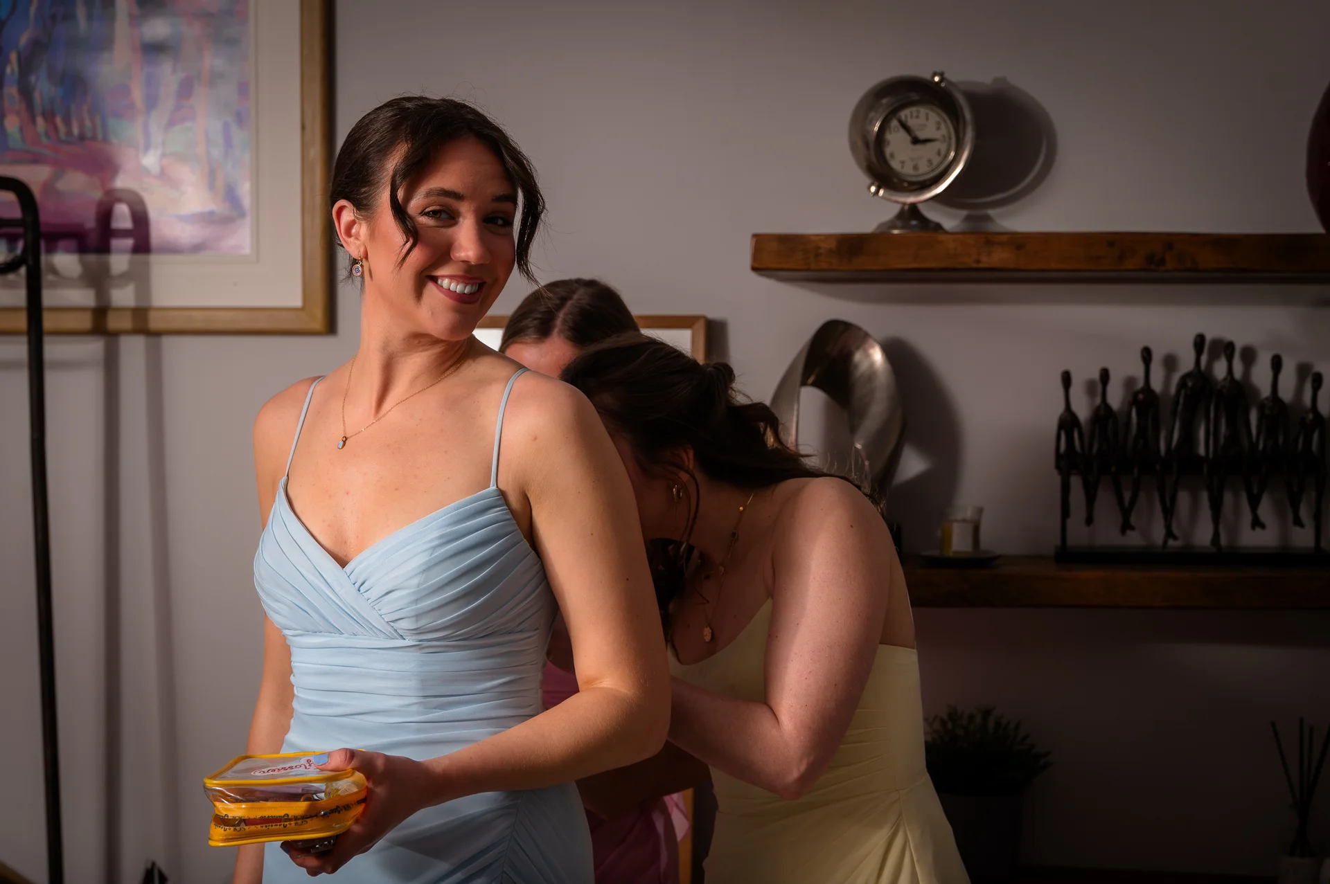 A smiling bridesmaid in a light blue dress with a ruched bodice stands in the foreground while another bridesmaid helps her prepare indoors.