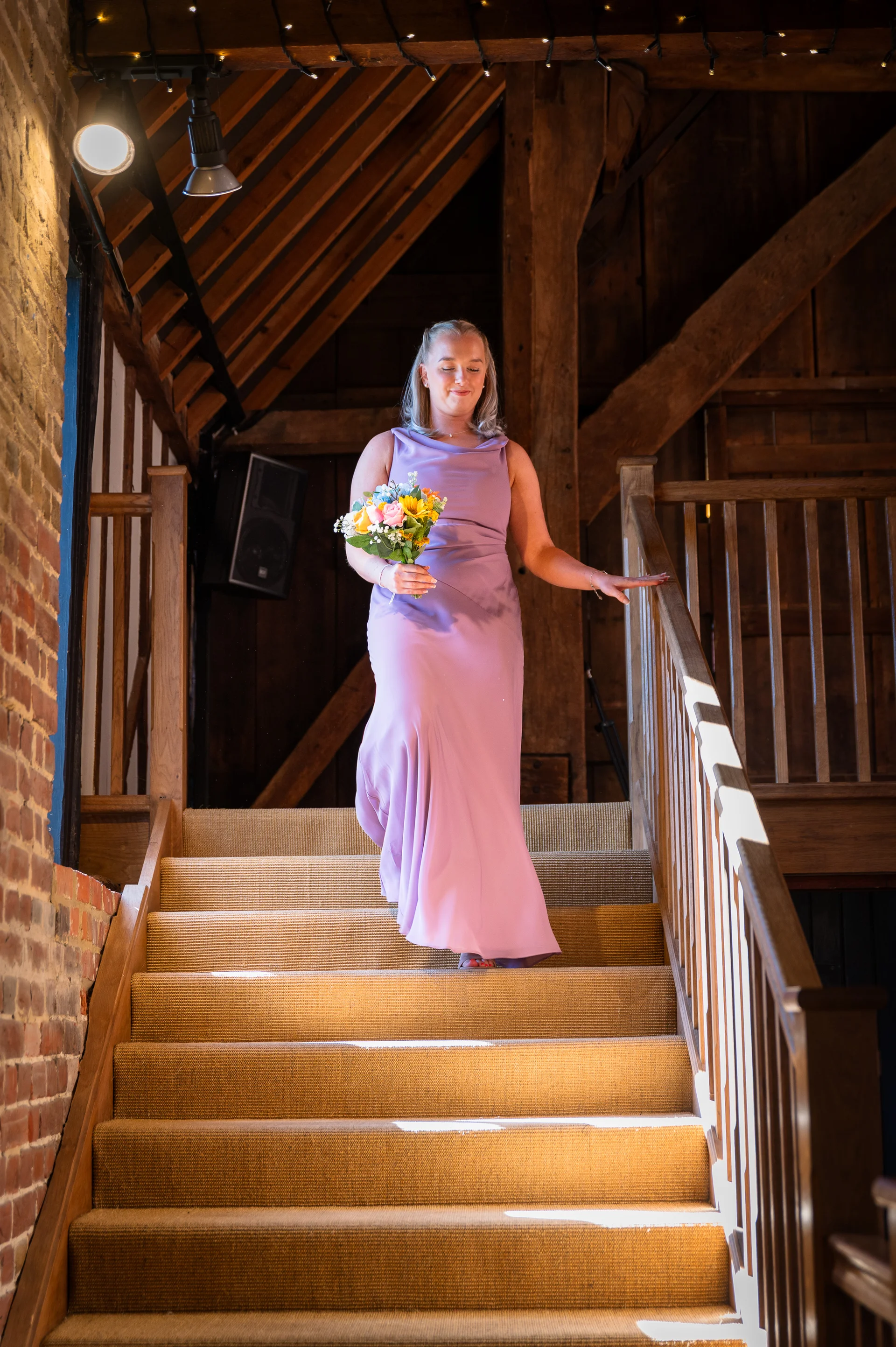 A bridesmaid in a sleek pink dress descends a wooden staircase inside an oak-framed barn, holding a colourful bouquet while sunlight streams through.