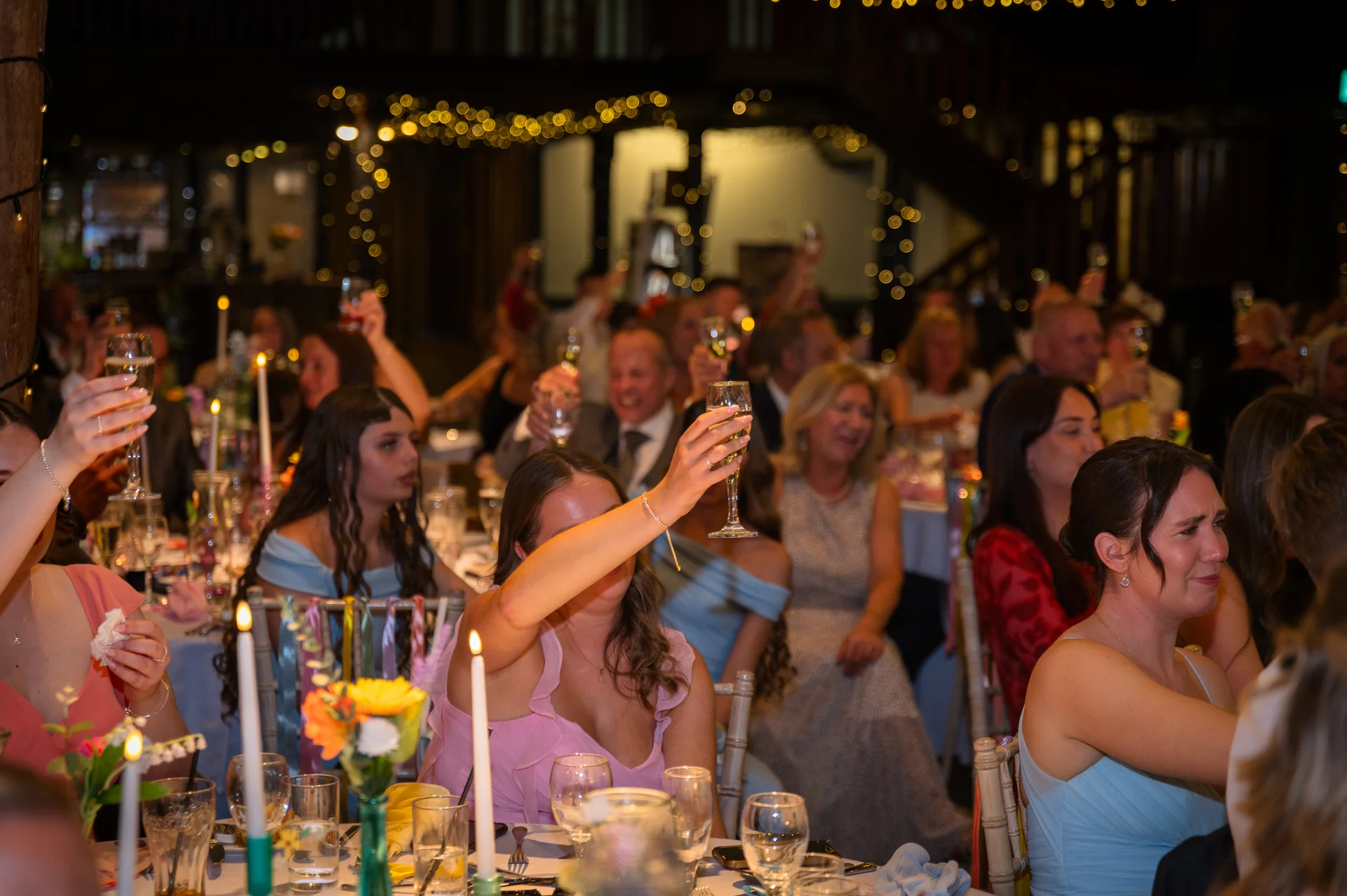 Wedding guests raise their champagne glasses in a toast during the speeches in a candlelit reception venue.