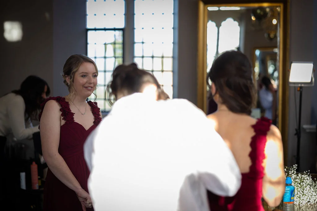 Bridesmaids in matching burgundy dresses share a joyful moment together while getting ready in a sunlit room.