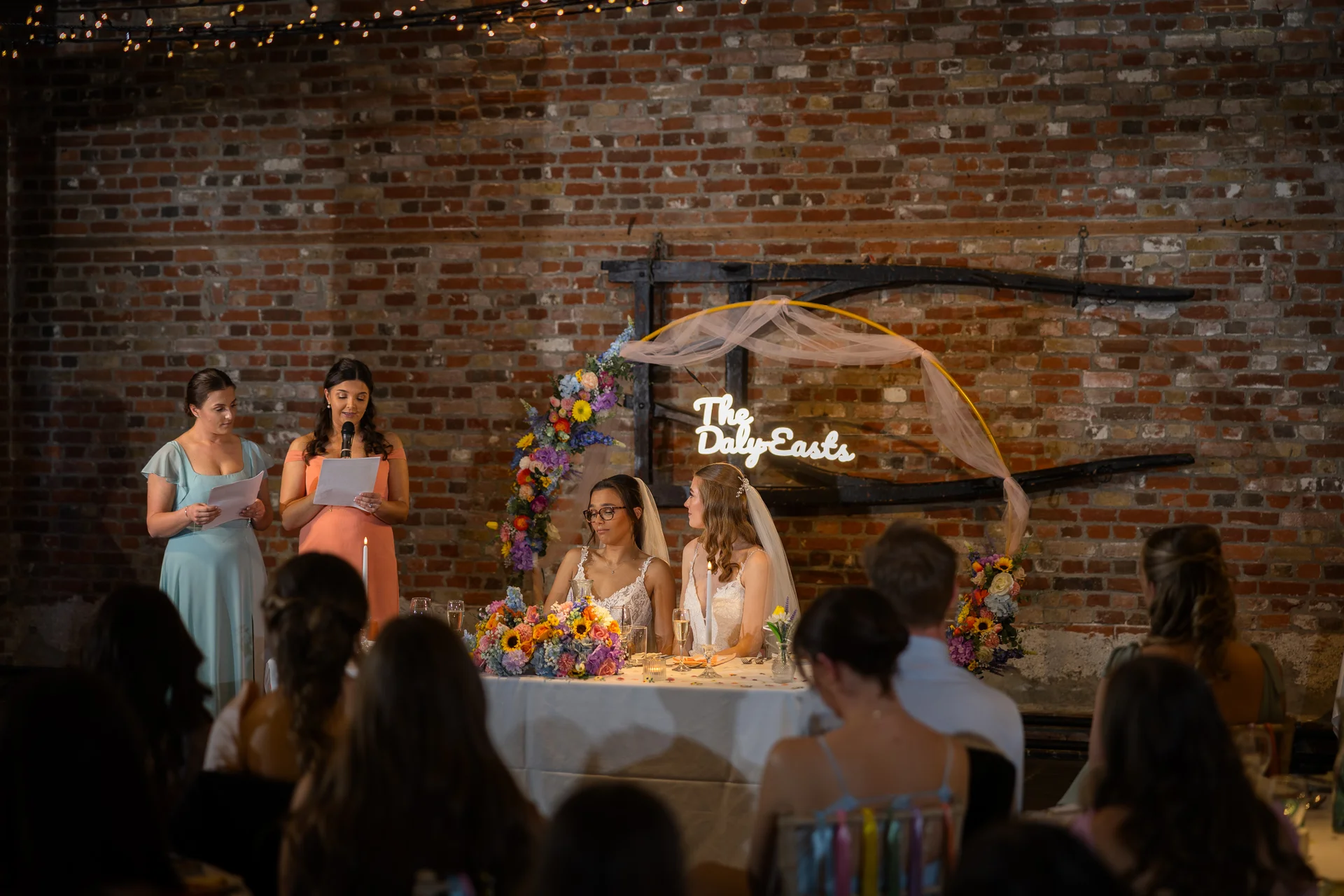 Two brides are seated at the top table during wedding speeches in a rustic warehouse venue with exposed brick walls, while bridesmaids stand nearby reading from notes.