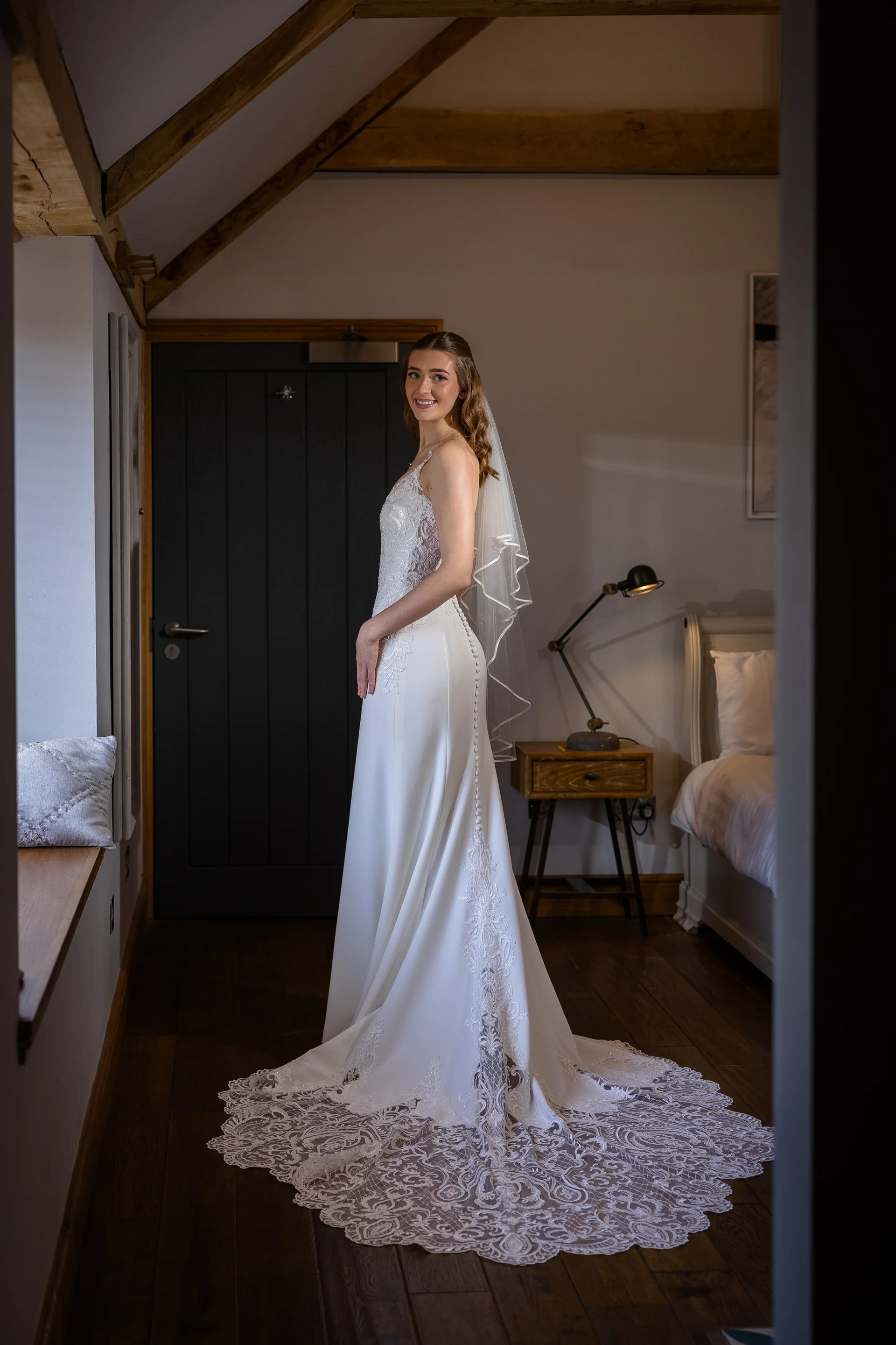 A stunning bride in an a-line crepe wedding dress with a dramatic lace train poses in a rustic bedroom with exposed wooden beams.