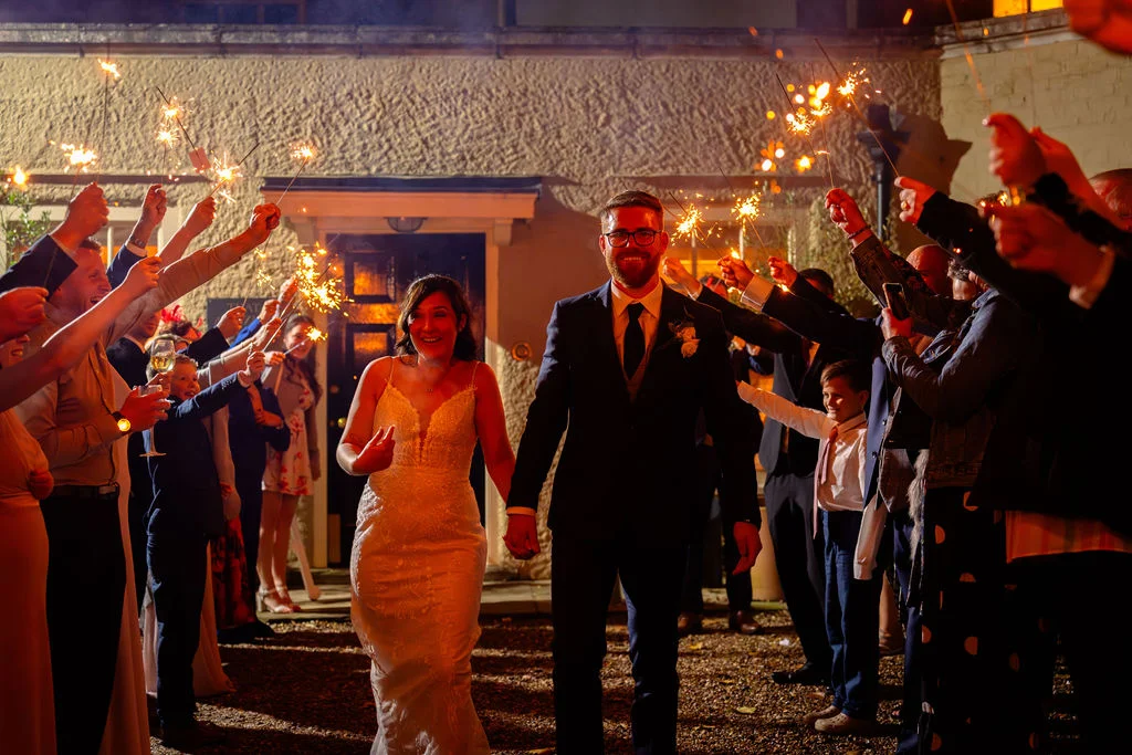 Newlyweds joyfully walk hand in hand through a glowing tunnel of sparklers held by cheering wedding guests at night.