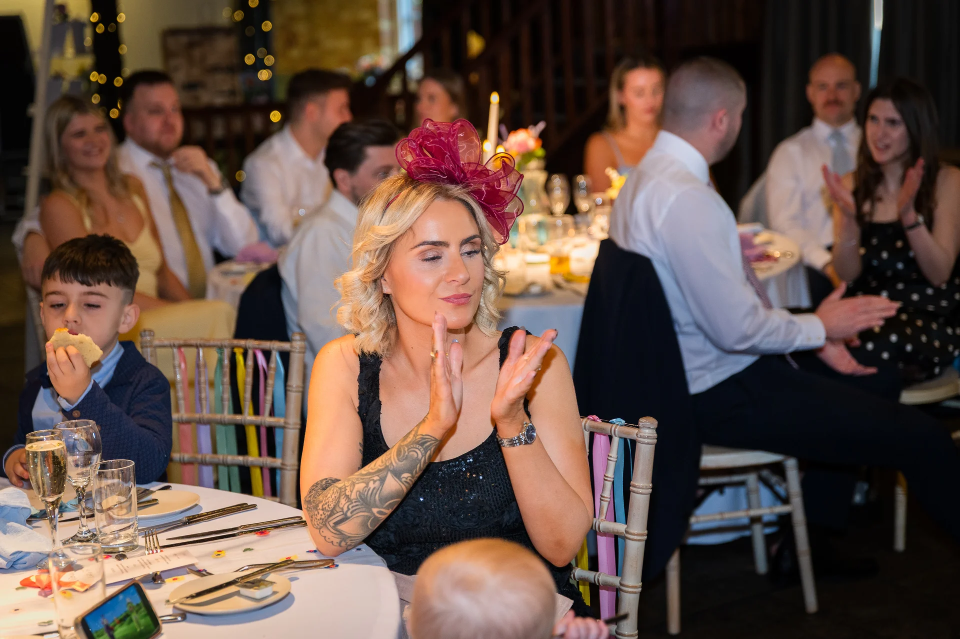 A wedding guest wearing a burgundy fascinator and a patterned black top applauds at the reception while sitting at a table with family and friends.