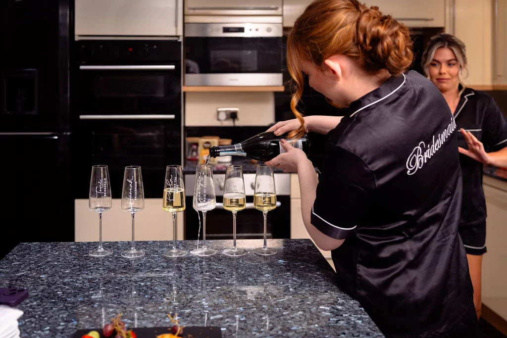 A bridesmaid in a black satin pyjama set pours champagne into personalised flutes lined up on a granite kitchen island while another bridesmaid watches.