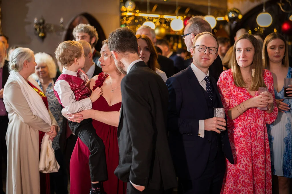 Candid photograph of wedding guests mingling and chatting during a lively indoor drinks reception with warm ambient lighting.