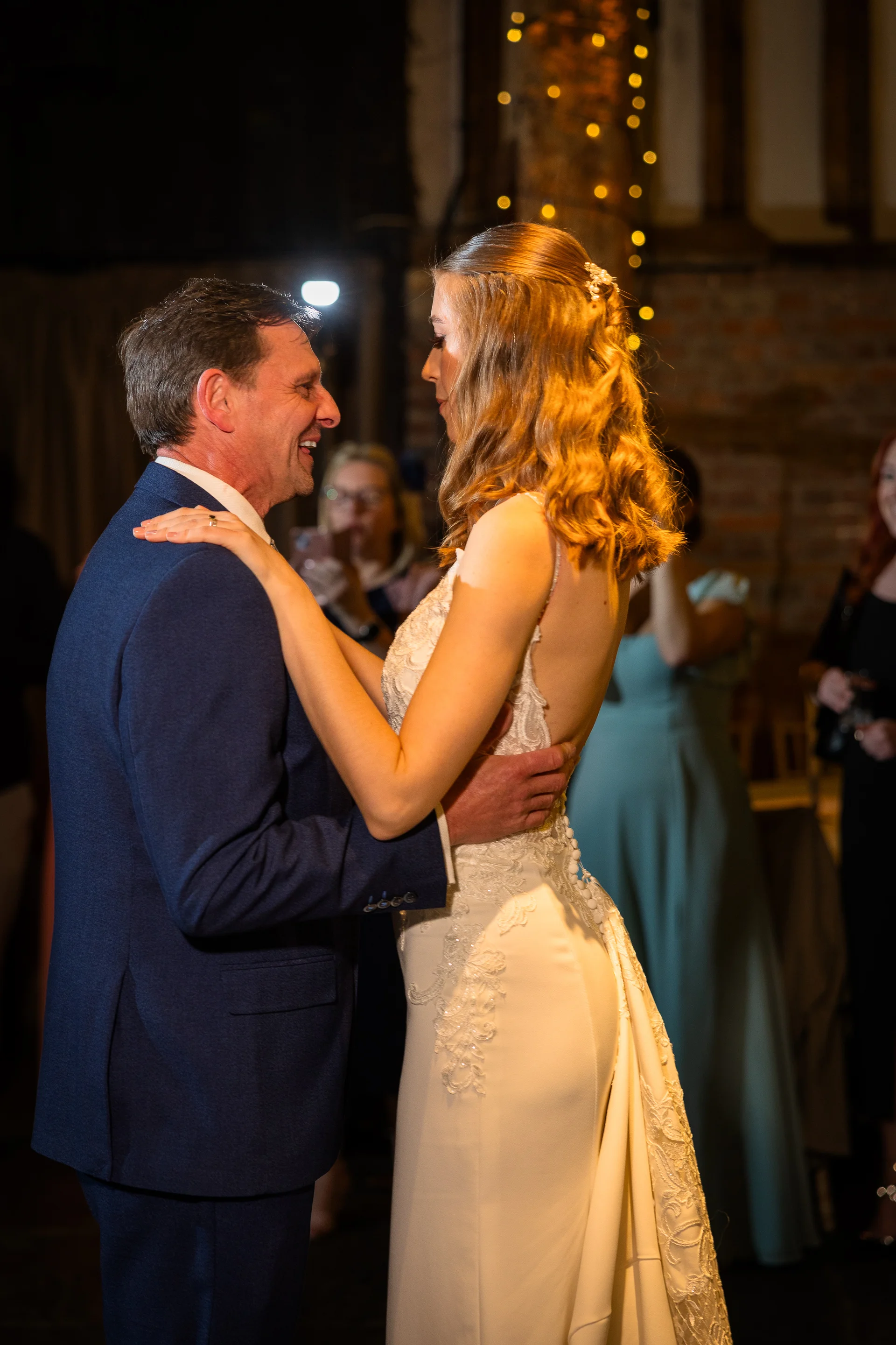 A smiling father and his bride daughter share a joyful father-daughter dance inside a rustic barn venue decorated with fairy lights.