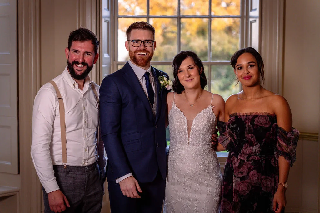 A happy wedding party poses indoors in front of a large window with autumn foliage visible outside, featuring a bride in a lace gown and groom in a navy suit.