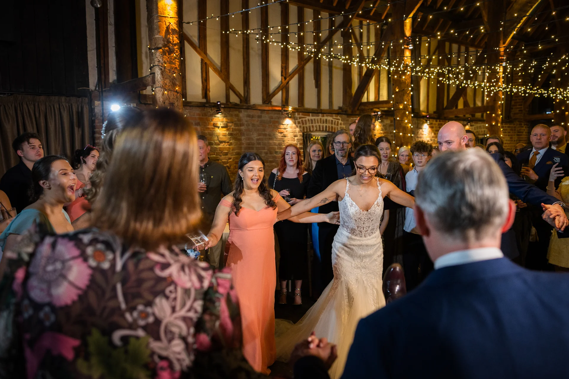 A joyful bride in a lace gown and glasses holds hands with guests forming a large dance circle inside a rustic barn illuminated by fairy lights.