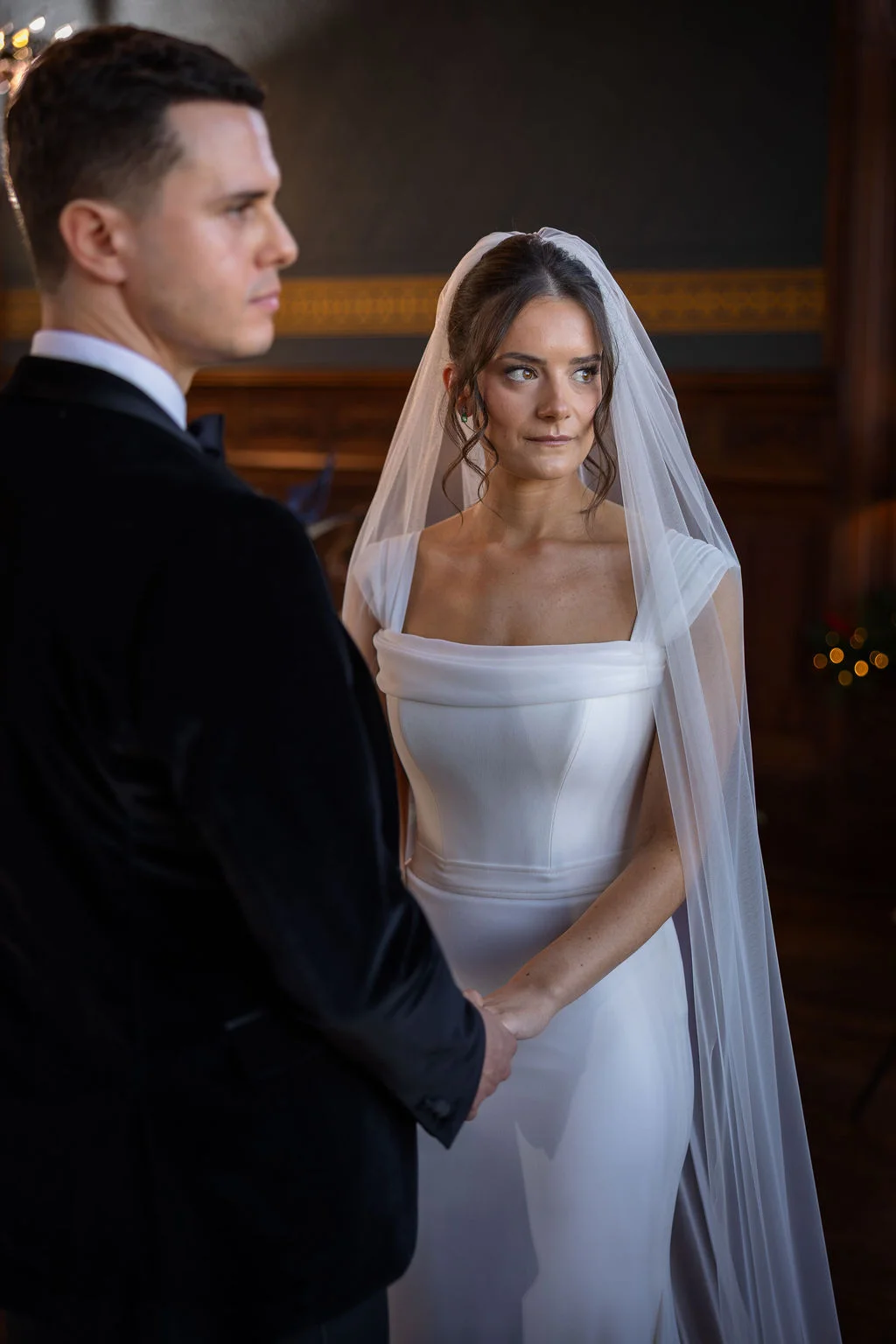 A newlywed bride wearing a chic square-neck gown and veil holds hands and shares a quiet moment with her groom in a black tuxedo.