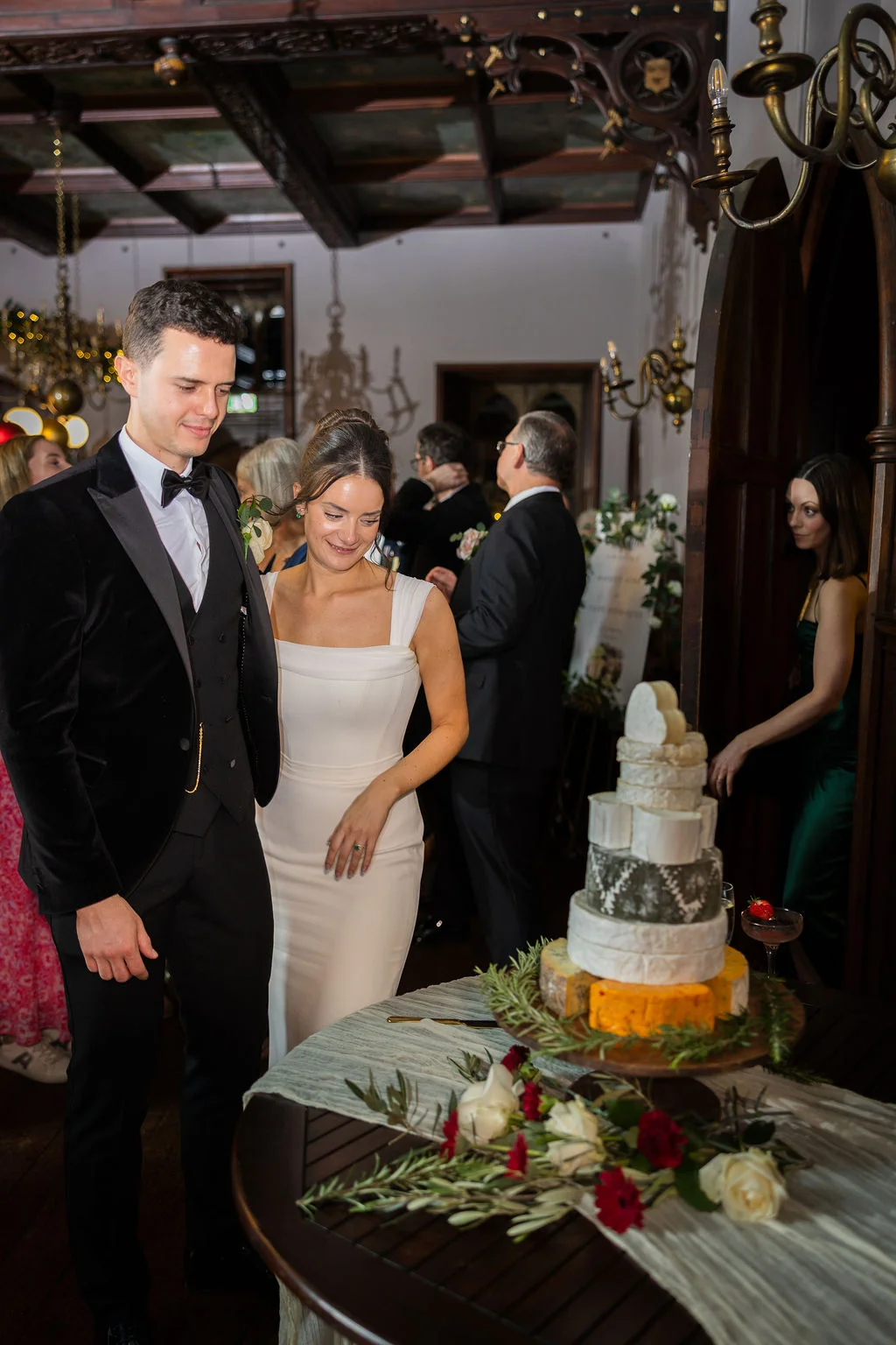 A newlywed couple stands together smiling next to a tiered cheese wheel wedding cake at an indoor evening reception inside a historic venue.